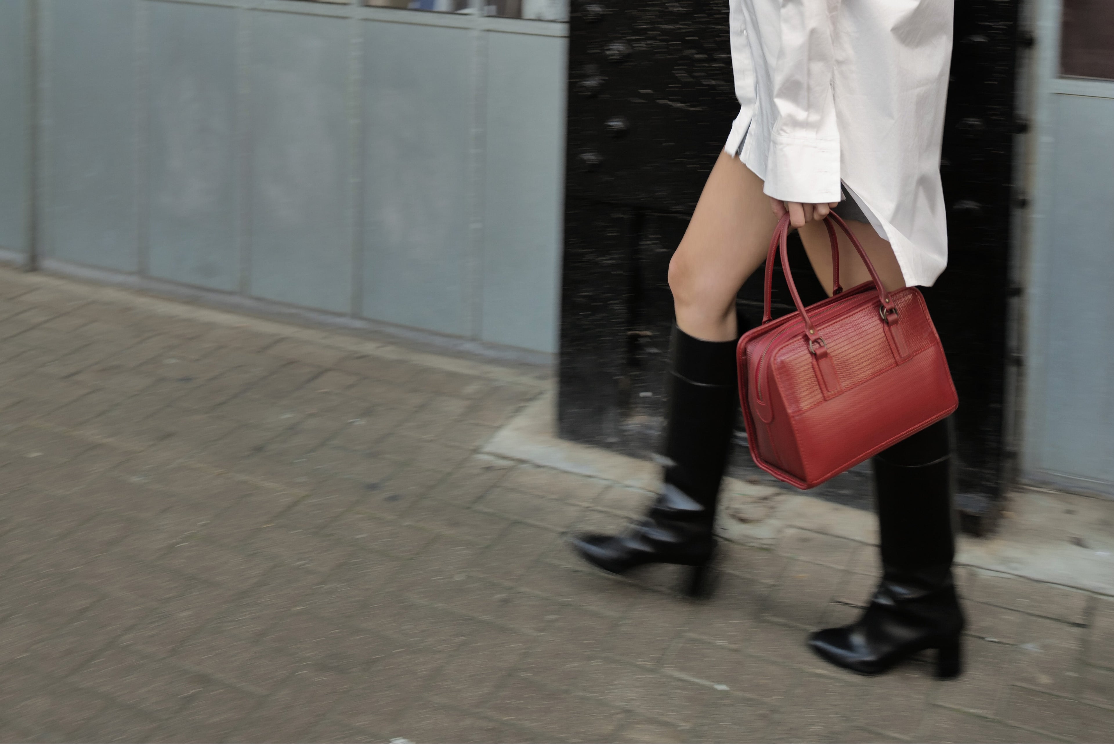 Person wearing black knee-high boots and holding a red handbag on a sidewalk.